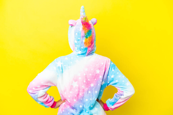 Girl with curly hair wearing a unicorn pajama isolated on yellow background in back position - Photo, image