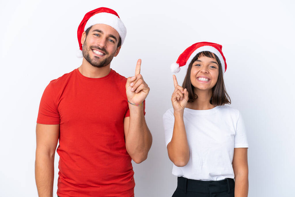 Beyaz arka planda izole edilmiş Noel şapkalı genç çift en iyinin işareti olarak parmağını kaldırıyor. - Fotoğraf, Görsel