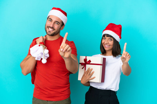Молода пара з різдвяним капелюхом роздає подарунки ізольовані на синьому фоні, показуючи і піднімаючи палець
 - Фото, зображення