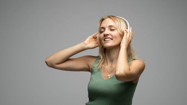 Beautiful attractive young blond woman wearing green t-shirt and glasses in white headphones listening music and smiling on grey background in studio. Relaxing and enjoying. Lifestyle. - Фото, изображение