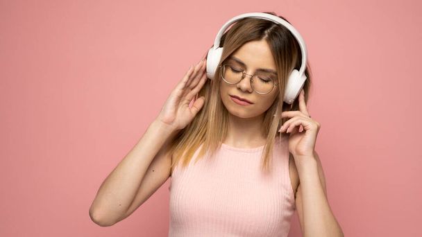 Beautiful attractive young blond woman wearing pink t-shirt and glasses in white headphones listening music and smiling on pink background in studio. Relaxing and enjoying. Lifestyle. - Photo, image