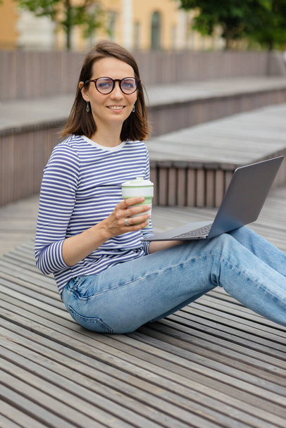 young and confident woman blogger or marketer drinking coffee and working on her laptop. work in a park or public place - Фото, зображення