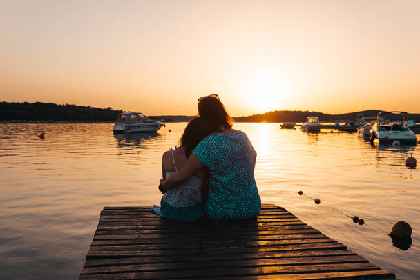 Mamá e hija sentadas en un muelle de madera, abrazándose unas a otras admirando el sol poniente sobre el mar - Foto, imagen