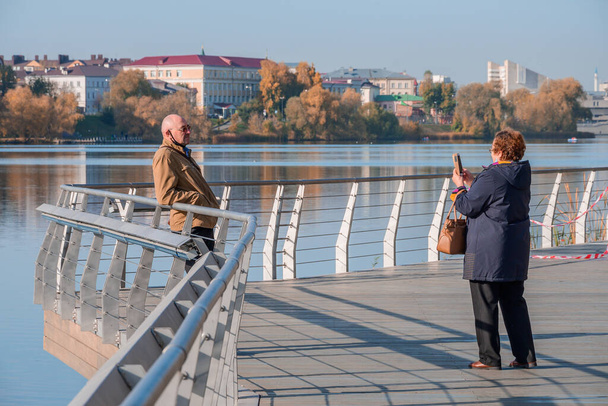 Kazan, Russia - October 14, 2020: An elderly couple taking pictures of each other on the phone on the wooden embankment of the city lake against the backdrop of the urban landscape on a sunny autumn day. - Photo, image