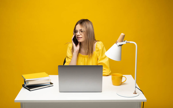 Young attractive business woman with a blond hair sitting at the table and working on a laptop and talking with a client on the phone. Young cheerful student girl talking with a smartphone. Studying. - Foto, afbeelding