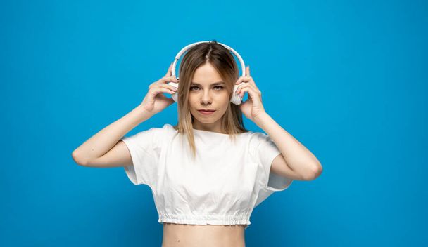 Beautiful attractive young blond woman wearing white t-shirt and glasses in white headphones listening music and smiling on blue background in studio. Relaxing and enjoying. Lifestyle. - Fotografie, Obrázek