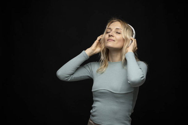 Beautiful attractive young blond woman wearing blue t-shirt and glasses in white headphones listening music and smiling on black background in studio. Relaxing and enjoying. Lifestyle. - Φωτογραφία, εικόνα