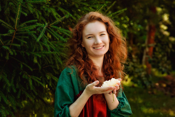 Portret rudej kobiety w parku. Kędzierzawa kobieta w pobliżu zielonych gałązek jodły trzyma małe płatki róży w dłoniach. - Zdjęcie, obraz