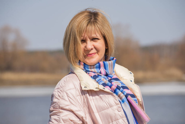 Een vrouw staat aan de oever van de rivier met een jas en een sjaal om haar nek. Volledig gezichtszicht. Kort kapsel blond. Tegen de achtergrond van de oever van het stuwmeer. Inheemse glimlach. - Foto, afbeelding