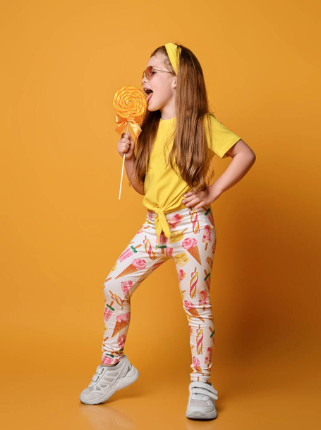 Happy redhead kid girl in yellow t-shirt, headband, colorful leggings and sneakers stands sideways licking big lollipop - Fotó, kép