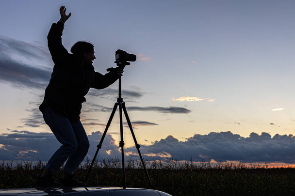 Uma menina fotografa o pôr do sol de um tripé, em pé no telhado de um carro no campo, espaço de cópia. - Foto, Imagem