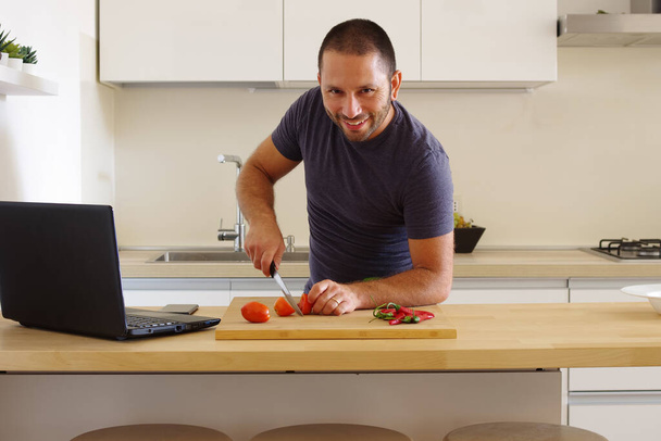 A white man cutting vegetables on a board next to his laptop in the kitchen - Fotoğraf, Görsel