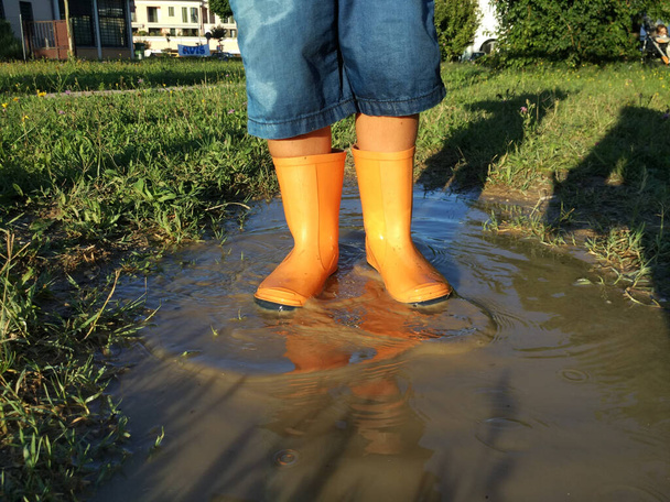 A vertical shot of a child's feet in orange rubber boots in muddy water surrounded by grass - Фото, зображення