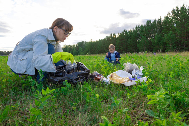 Μαμά με λαστιχένια γάντια με το γιο της στη φύση συλλέγουν πλαστικά σκουπίδια σε μια ηλιόλουστη μέρα του καλοκαιριού στο φόντο των δέντρων. Προστασία του περιβάλλοντος. Επιλεκτική εστίαση - Φωτογραφία, εικόνα