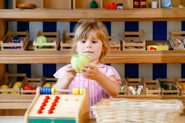 Mała przedszkolanka bawi się jedzeniem i zakupami drewnianych zabawek. Szczęśliwy aktywne dziecko gra roli kasjera lub sprzedawcy, w sklepie z drewnem lub supermarkecie. Edukacja, zajęcia dla dzieci. - Zdjęcie, obraz