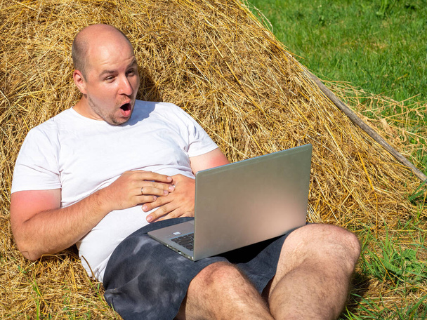 Tişört ve şort giymiş orta yaşlı bir adam elinde dizüstü bilgisayarla sarı bir samanlıkta yatıyor. Uzaktan çalışma, eğitim. Kırsal bölge. Adamın yüzünde bir sürpriz, duygular... - Fotoğraf, Görsel