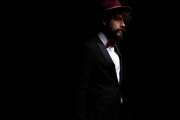 vista lateral de un atractivo hombre de negocios mirando hacia otro lado y llevando un sombrero de color burdeos sobre fondo negro - Foto, imagen