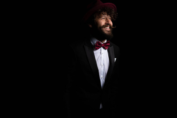 sexy businessman smiling wide and wearing a burgundy hat on black studio background - Фото, изображение