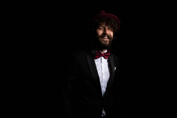 giovane uomo d'affari sorridente alla fotocamera e indossa un cappello bordeaux sullo sfondo nero - Foto, immagini