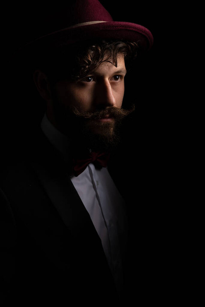 portrait d'un jeune homme d'affaires détournant les yeux, coiffé d'un chapeau bordeaux sur fond noir - Photo, image