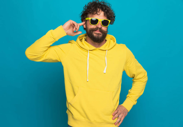 portrait de jeune homme heureux à capuche jaune pointant du doigt à l'oreille et souriant tout en se tenant debout et posant sur fond bleu en studio - Photo, image