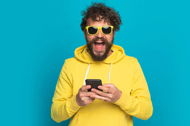 happy young man with sunglasses and yellow hoodie holding phone, reading news and laughing on blue background in studio - Photo, Image
