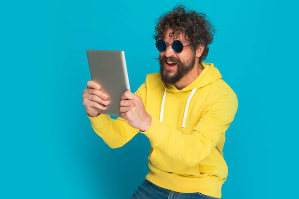 portré lelkes szakállas férfi sárga kapucnis retro szemüveg tartó tab, nevetés és pózol a kék háttér stúdió - Fotó, kép