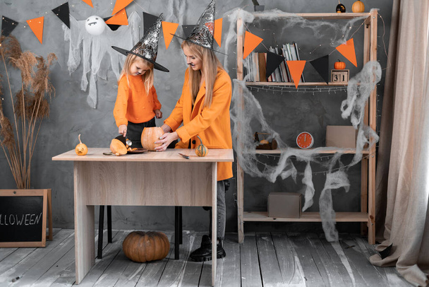 Wiedźma mama i córka przygotowują dynię w domu na Halloween. Dziewczyna wyjmuje nasiona i miąższ pomarańczy. Dziecko z mamą w kapeluszach czarownic zrobić jack na latarni. - Zdjęcie, obraz