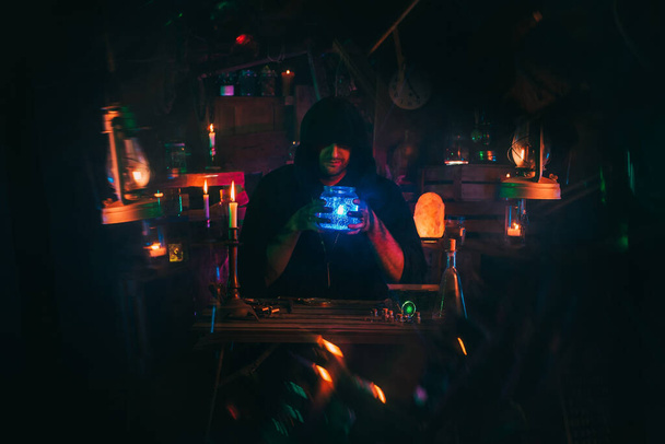 tovenaar alchemist in donkere kleren is bezig met toverdrankje maken in een ambachtelijke werkplaats - Foto, afbeelding