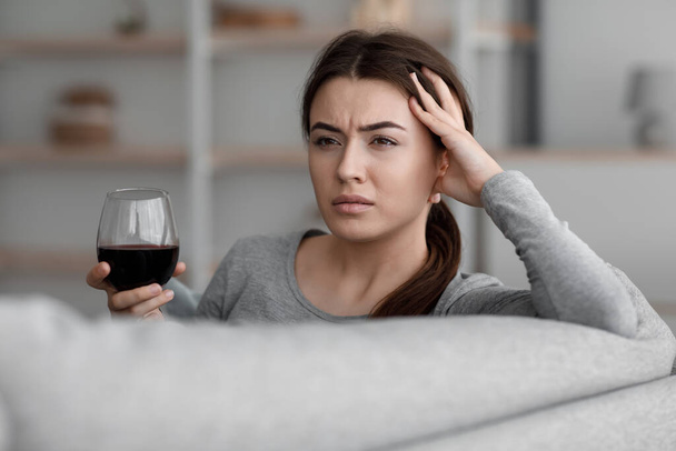 Μεθυσμένη δυστυχισμένη Καυκάσια νεαρή γυναίκα με ένα ποτήρι κρασί που υποφέρει από κατάθλιψη και πονοκέφαλο και χαλαρώνει στο σπίτι - Φωτογραφία, εικόνα