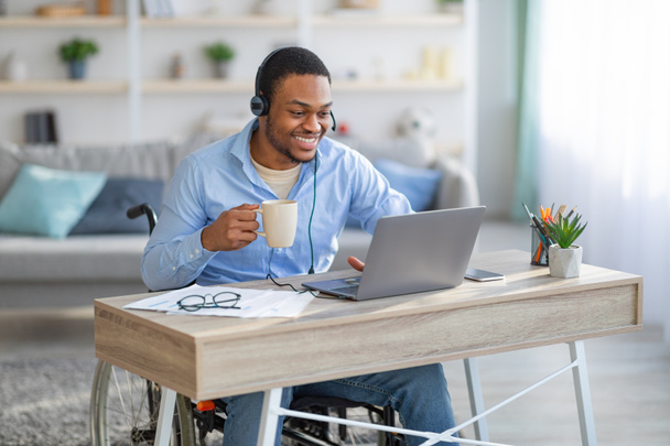 Remote job for disabled people. Black guy in wheelchair wearing headset, using laptop, drinking coffee at home office - Photo, Image