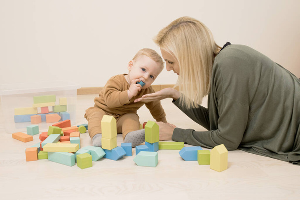 Child plays with wooden toys on the floor. Baby has part of toy in his mouth, mother held out her hand to pick it up. Concept of early development, toddler takes toys in his mouth, danger of choking - Фото, изображение