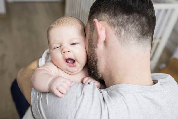 Newborn baby yawns in the hands of the father. Dad takes care of his newborn son at home. Concept of  toddler health care, parenting, family, fatherhood, sleepy baby, motion sickness, baby care. - 写真・画像
