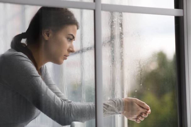 Депресивна сумна біла тисячолітня жінка страждає від стресу і дивиться у вікно вдома
 - Фото, зображення