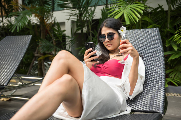 Asyalı, kırmızı elbiseli, tek parça mayolu genç bir kadın telefon kullanıyor ve havuzda yatak sandalyesinde domates suyu içiyor. Bayan kız, yaz tatilinde lüks otelde dinleniyor.. - Fotoğraf, Görsel