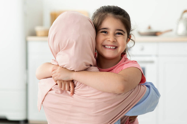 Ευτυχισμένο κοριτσάκι που αγκαλιάζει τη μουσουλμάνα μαμά της με το χιτζάμπ στο σπίτι, κοντινό πλάνο - Φωτογραφία, εικόνα