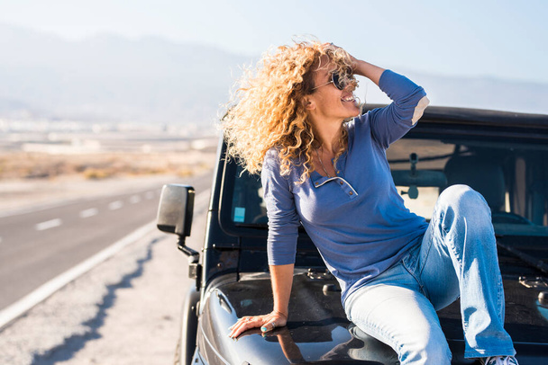 Happy beautiful mature woman in sunglasses posing with hand in curly hair while sitting on bonnet of jeep at highway - Photo, Image