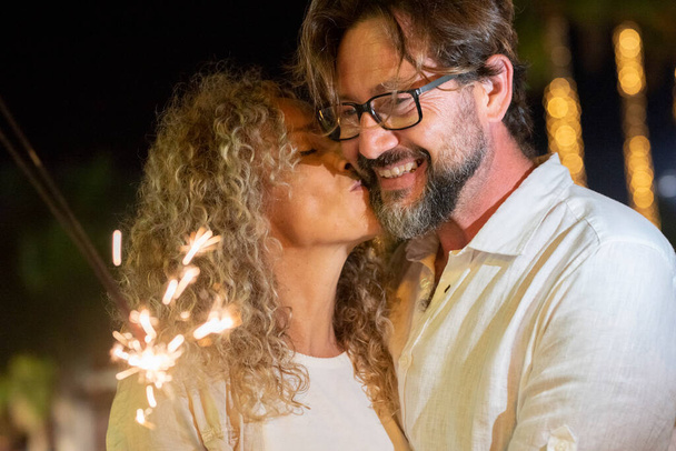 Beautiful mature woman kissing to cheerful man looking at burning sparkle - Photo, Image