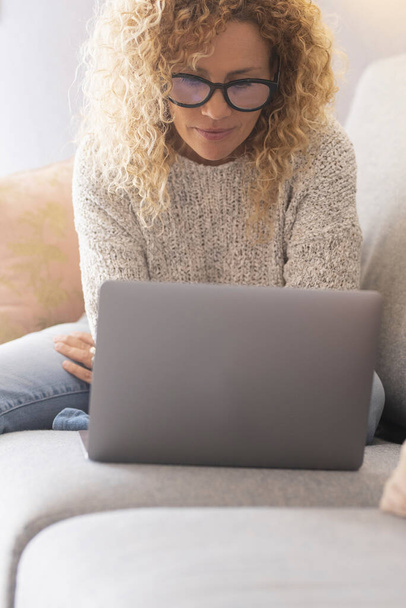 Зрелая женщина с кудрявыми волосами в очках сидит на диване, а дома пользуется ноутбуком - Фото, изображение