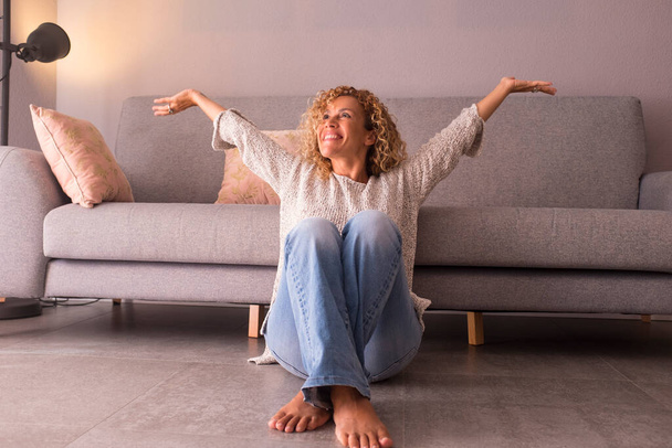 Cheerful young woman sitting on floor with arms raised at home.  - Foto, afbeelding