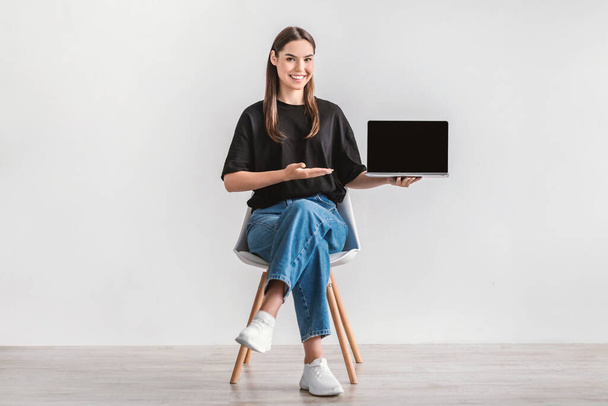 Positiivinen nuori nainen osoittaa kannettavan tietokoneen tyhjällä näytöllä, istuu tuolilla vastaan valkoinen studio seinään, mockup verkkosivuilla - Valokuva, kuva