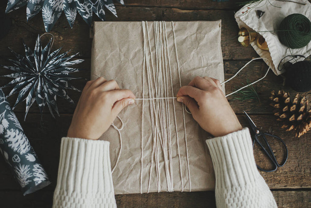 Hände wickeln stilvolles Weihnachtsgeschenk in Bastelpapier mit Schnur auf rustikalem Holztisch mit blauen Papiersternen. Flach lag er. Stimmungsvolles Stimmungsbild, nordischer Stil. Frohe Weihnachten und frohe Feiertage! - Foto, Bild