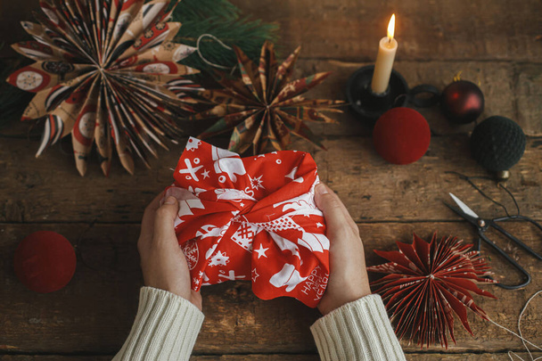 Prezent świąteczny Furoshiki. Dłonie gospodarstwa prezent świąteczny zawinięty w czerwony świąteczny materiał na rustykalnym drewnianym stole z nożyczkami, gwiazda papieru, świeca, ozdoby. Atmosferyczny czas nastrojów. Zero odpadów wakacyjnych - Zdjęcie, obraz