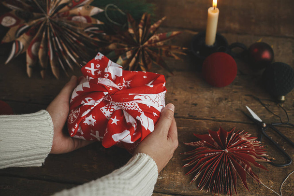 Furoshiki Weihnachtsgeschenk. Hände halten Weihnachtsgeschenk in rotem Feststoff auf rustikalem Holztisch mit Schere, Papierstern, Kerze, Ornamenten. Stimmungsvolle Zeit. Zero Waste Urlaub - Foto, Bild