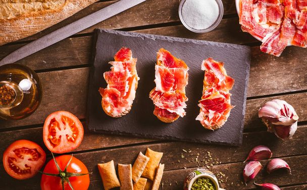 Iberian ham toasts on a slate plate on a brown wooden table with some ingredientes around it. Spanish typical food. Pork meat. - Photo, Image