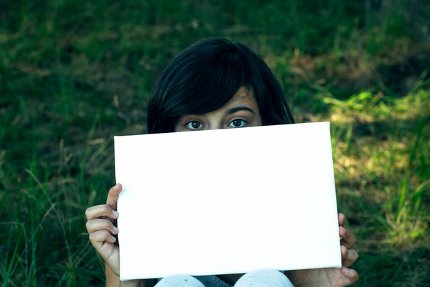 Κορίτσι που κρατά καθαρό λευκό χαρτί (banner για το μήνυμα), στο καλοκαιρινό πάρκο. - Φωτογραφία, εικόνα