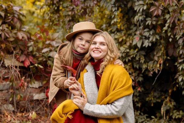 A young adult blonde mother with a 7-year-old daughter on a walk in the autumn park hug and laugh - Foto, imagen