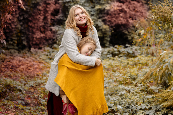 A young adult blonde mother with a 7-year-old daughter on a walk in the autumn park hug and laugh - Fotó, kép