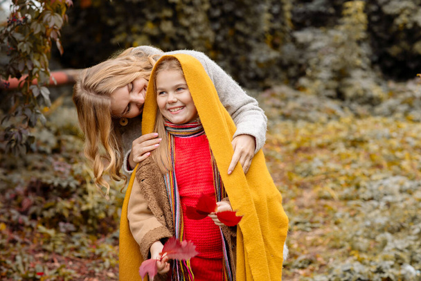 A young adult blonde mother with a 7-year-old daughter on a walk in the autumn park hug and laugh - Fotografie, Obrázek