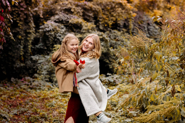 A young adult blonde mother with a 7-year-old daughter on a walk in the autumn park hug and laugh - Foto, afbeelding
